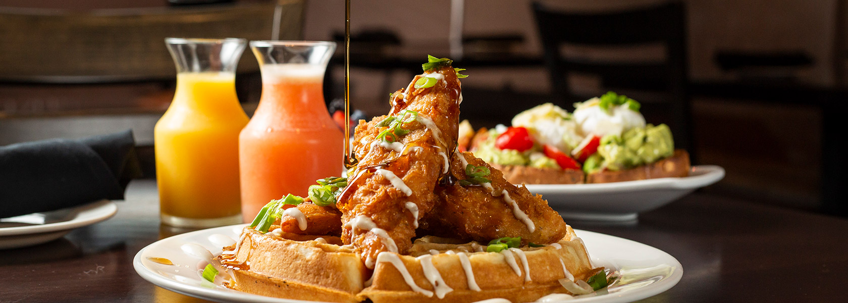 Syrup being drizzled over fried chicken on waffles with breakfast juices in background