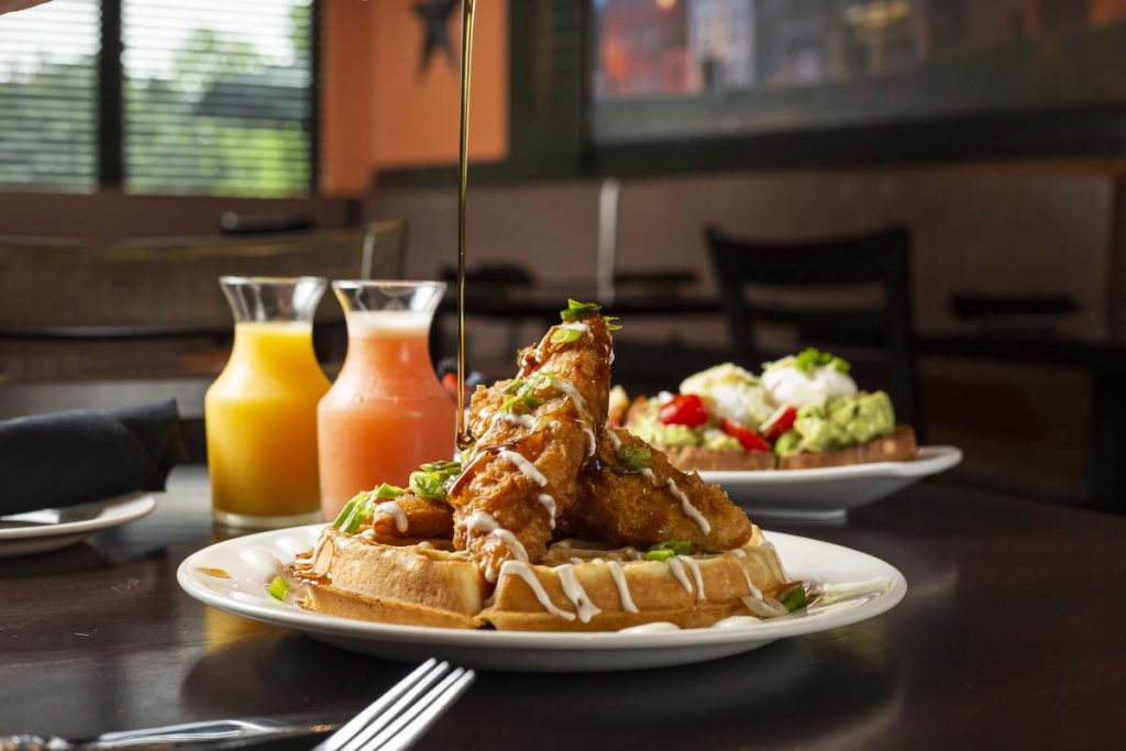 Syrup being drizzled over chicken and waffles with breakfast juices in background