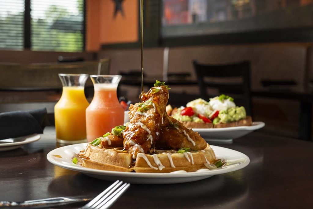 Syrup being drizzled over chicken and waffles with breakfast juices in background